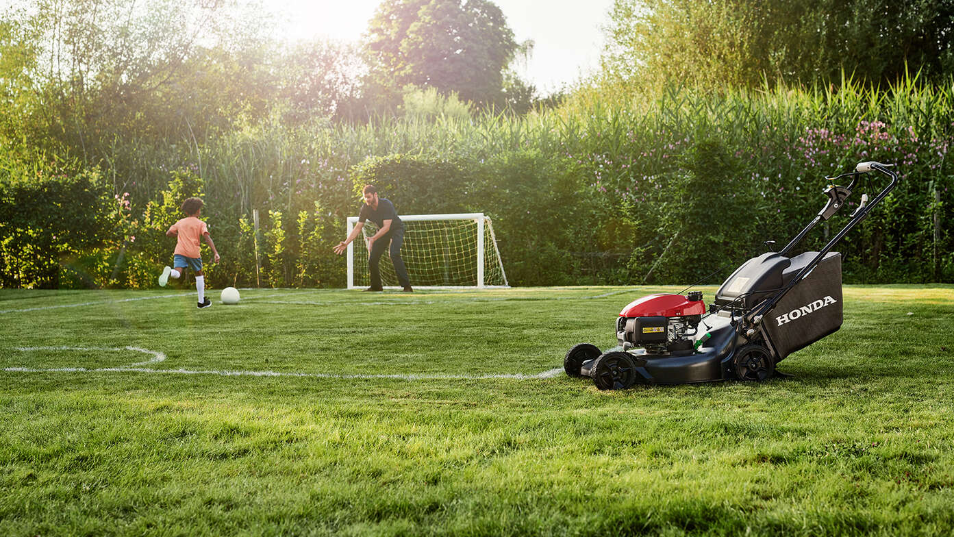 Muž a dieťa hrajú v záhrade futbal. Bočný pohľad na kosačku Honda HRN. 